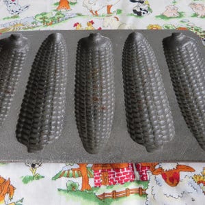 May include: A dark gray cast iron corn bread pan featuring five distinct, elongated molds shaped like ears of corn. The rectangular pan includes two integrated side handles, each with a circular opening, and a textured surface throughout.