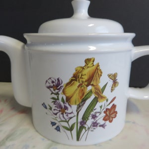 May include: White ceramic teapot with a floral design featuring a yellow iris, purple and white flowers, and a butterfly. The teapot has a lid and a handle.