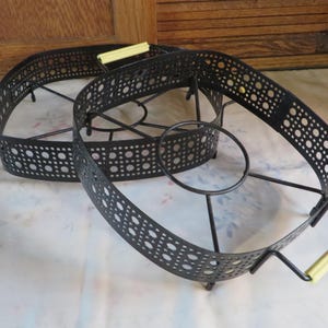 May include: Two black metal serving stands with oval frames and perforated designs. Each stand has a circular base and yellow handles. The stands are designed to hold serving dishes and are likely vintage.