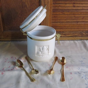 May include: A white ceramic tea canister with a gold rim and clasp, featuring a raised teapot design. Four gold-colored spoons are arranged in front of the canister. The canister's lid is slightly ajar.