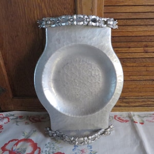 May include: A silver hammered metal serving tray with a scalloped edge and floral design handles. The tray has a hammered texture and a shiny finish.