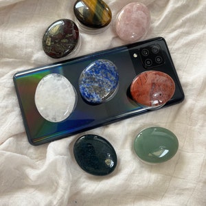 May include: Assortment of seven smooth, polished, oval-shaped stones in various colours, including green, black, white, red, pink, blue, and brown. The stones are arranged on a white fabric surface and a black smartphone.