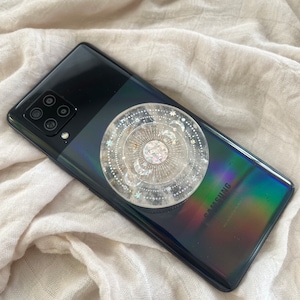 May include: A black Samsung smartphone with a clear phone grip featuring a celestial design with moons, stars, and a sun. The grip is attached to the back of the phone.