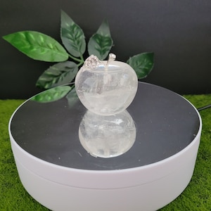 May include: A clear quartz crystal carved into the shape of an apple with a silver leaf and rhinestone accents. The apple is displayed on a white rotating platform with a mirrored surface.