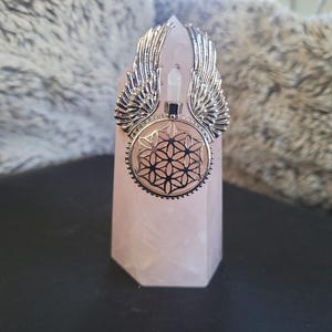May include: A silver pendant with wing detailing and a geometric pattern, displayed on a pink crystal. The pendant includes a clear crystal and a black square. The crystal is a pale pink colour.