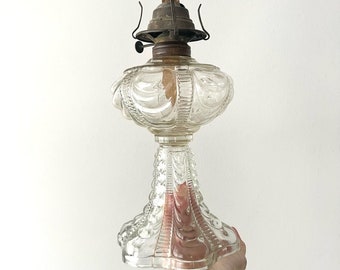 May include: A clear glass oil lamp with a decorative, ornate design. The lamp has a round, scalloped base and a clear glass chimney.