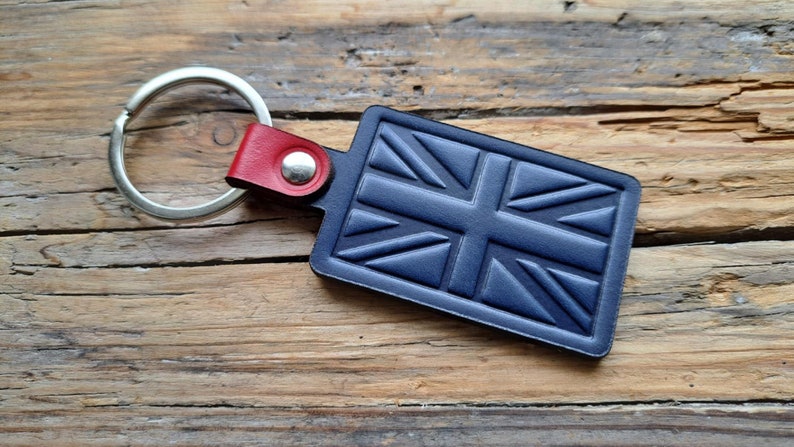 Puede incluir: Un llavero de cuero azul con un dise&ntilde;o de Union Jack y un anillo de llave plateado. El llavero tiene una correa de cuero roja con un remache plateado.