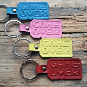 May include: Four leather keychains in blue, pink, yellow, and red, each with the words "GAME OVER" in pixelated font. The keychains have silver key rings and are arranged on a rustic wooden surface.