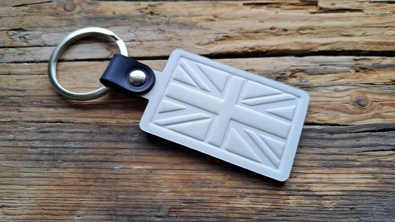 Puede incluir: Un llavero plateado con una correa de cuero y un dise&ntilde;o de Union Jack blanco en relieve.