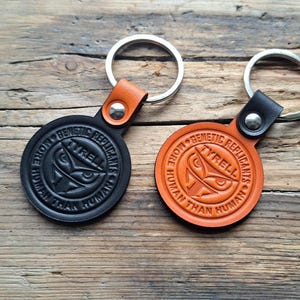 May include: Two leather keyrings with silver rings. One is black with an orange strap, the other is orange with a black strap. Both feature the text "GENETIC REPLICANTS MORE HUMAN THAN HUMAN" and an owl design.