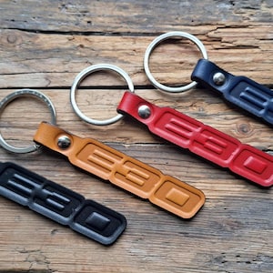 May include: Four leather keychains in black, tan, red, and navy blue, each with a silver keyring. The keychains are embossed with the letters "E30" and are arranged on a wooden surface.