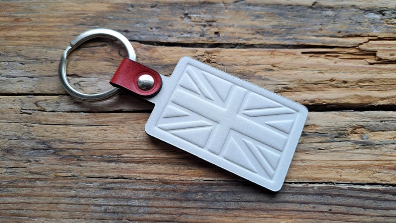Puede incluir: Llavero de cuero blanco con un anillo de llave plateado y una correa de cuero roja. El llavero presenta un dise&ntilde;o de Union Jack en relieve.