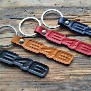 Pode incluir: Porta-chaves de couro preto, castanho, vermelho e azul, cada um com um porta-chaves prateado. Os porta-chaves têm a forma das letras "E46". Os porta-chaves estão dispostos numa superfície de madeira.