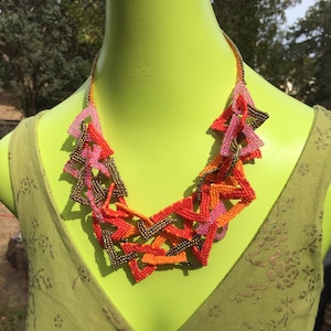May include: A multi-colored beaded necklace with a geometric design. The necklace is made of pink, orange, and gold beads and is arranged in a series of triangles. The necklace is displayed on a green mannequin.