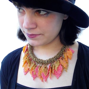May include: A woman wearing a black hat and a necklace made of beaded leaves in shades of pink and gold.
