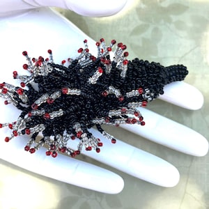 May include: A black beaded brooch with red and clear accents. The brooch is made of small beads, clustered together to create a spiky, textured design. The brooch is held in a gloved hand.