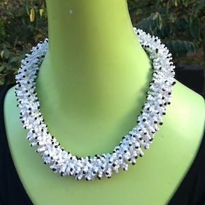 Pode incluir: Um colar de contas brancas e pretas com um design espesso e texturizado. O colar é feito de contas pequenas e redondas que são dispostas em um padrão espiral.