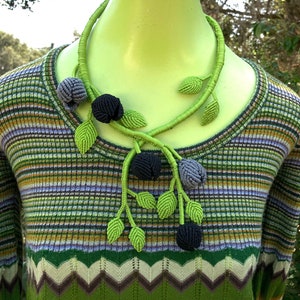 May include: A green crocheted necklace with black and gray flower accents. The necklace is made of multiple strands of yarn and features a leafy design.