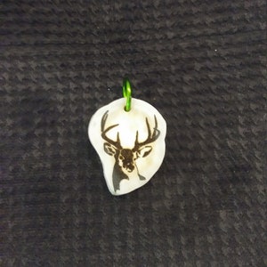 May include: A white ceramic pendant with a green metal loop. The pendant features a black silhouette of a deer head with antlers.