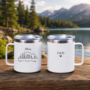 May include: Two white stainless steel mugs with clear lids and handles. The left mug features a black line drawing of mountains, trees, and a camper with the text Olivia and Live Love Camp. The right mug displays the letters O&M with a small heart.