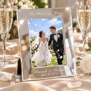 Op de afbeelding: Een rechthoekige zilverkleurige metalen fotolijst met een gepolijste afwerking. De bovenrand is gegraveerd met de tekst The Wedding Day en de onderrand bevat de gegraveerde tekst Hannah & Thoman Est. 2026, met een trouwfoto erin.
