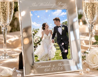 Puede incluir: Un marco de fotos rectangular de metal plateado con acabado pulido. El borde superior está grabado con el texto The Wedding Day, y el borde inferior presenta el texto grabado Hannah & Thoman Est. 2026, conteniendo una foto de boda.