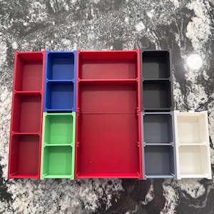 May include: A collection of colorful, small, modular shelves in red, blue, green, black, gray, and white. Each shelf unit has a rectangular shape with two or three compartments, suitable for organizing small items. The shelves are arranged on a speckled surface.