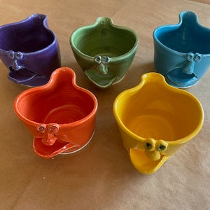May include: Five ceramic mugs with whimsical faces. The mugs are in various colors, including purple, green, blue, orange, and yellow. Each mug has a unique face with eyes, a mouth, and a nose.