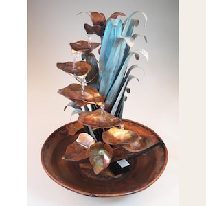 May include: A copper tabletop fountain with a cascading water feature. The fountain is shaped like a plant with copper leaves and blue-green stems. The water flows from the top leaves down to the base of the fountain.