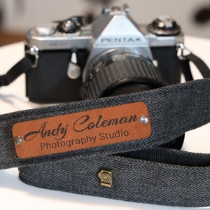 May include: A black and gray camera strap with a brown leather patch that says "Andy Coleman Photography Studio".