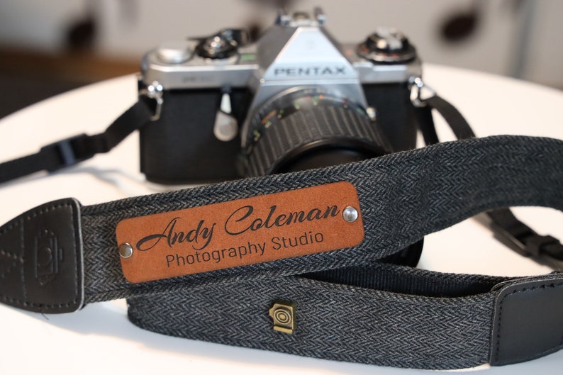 May include: A black and gray camera strap with a brown leather patch that says "Andy Coleman Photography Studio".