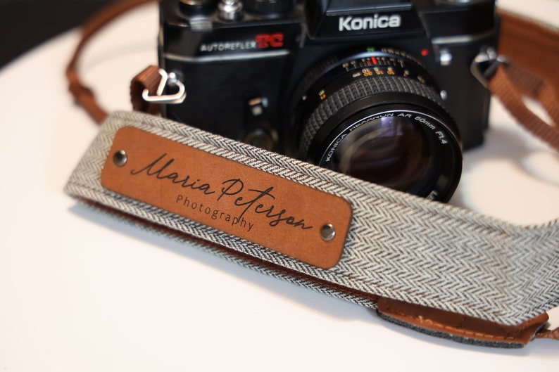 May include: A brown leather camera strap with a white and gray herringbone pattern. The strap has a brown leather patch with the text "Maria Peterson Photography" embossed in black.