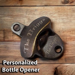 May include: A personalized bottle opener with a dark metal finish. The opener has a ring on the top and the text "The Briggs" is engraved on the front and back.