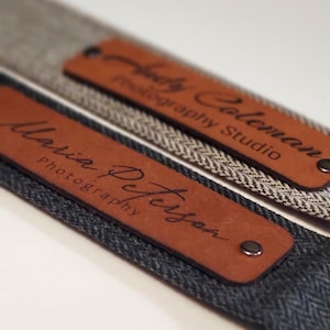 May include: Two brown leather camera straps with metal rivets. The straps are personalized with the names "Maria Peterson Photography" and "Holly Coleman Photography Studio".