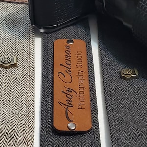 May include: Three camera straps with personalized leather name tags. The straps are made of a herringbone fabric in gray, white, and black. The name tags read "Maria Peterson Photography", "Andy Coleman Photography Studio", and a third tag that is not visible.