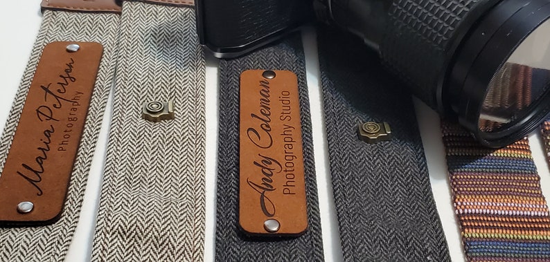 May include: Three camera straps with personalized leather name tags. The straps are made of a herringbone fabric in gray, white, and black. The name tags read "Maria Peterson Photography", "Andy Coleman Photography Studio", and a third tag that is not visible.