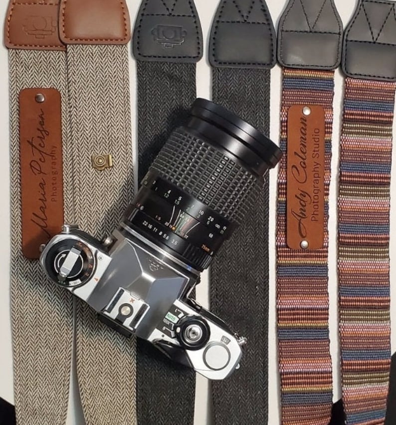 May include: Four camera straps with different patterns and colors. The straps are made of fabric and leather. The straps have metal buckles and are adjustable. The straps have personalized name tags with the text "Maria Peterson Photography" and "Andy Coleman Photography Studio".