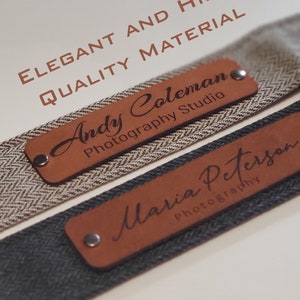 May include: Two brown leather camera straps with engraved text. The top strap says "Andy Coleman Photography Studio" and the bottom strap says "Maria Peterson Photography". The straps are made of high quality material.