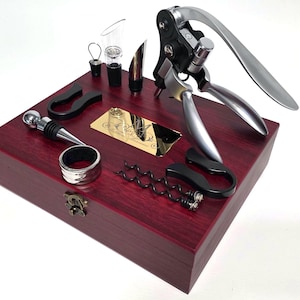 Könnte beinhalten: Ein Weinöffner-Set mit verschiedenen Werkzeugen, darunter ein Korkenzieher, ein Flaschenverschluss und ein Ausgießer, angeordnet auf einer dunkelroten Holzkiste. Das Set enthält einen silber-schwarzen Hebelkorkenzieher.