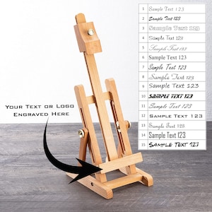 Könnte beinhalten: Ein kleiner, hölzerner Künstlerstaffelei mit verstellbarer Höhe und Neigung. Die Staffelei ist aus hellem Holz gefertigt und hat messingfarbene Beschläge. Der Text "YOUR TEXT OR LOGO ENGRAVED HERE" ist sichtbar. Eine Tabelle mit dem Text "Sample Text 123" ist ebenfalls vorhanden.