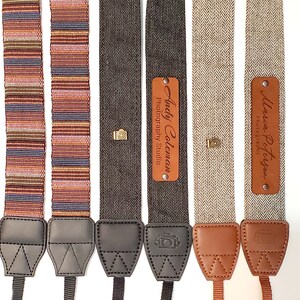 May include: Four camera straps with personalized leather name tags. The straps are made of different fabrics, including a striped fabric, a herringbone fabric, and a plain fabric. The name tags are brown leather with gold lettering.