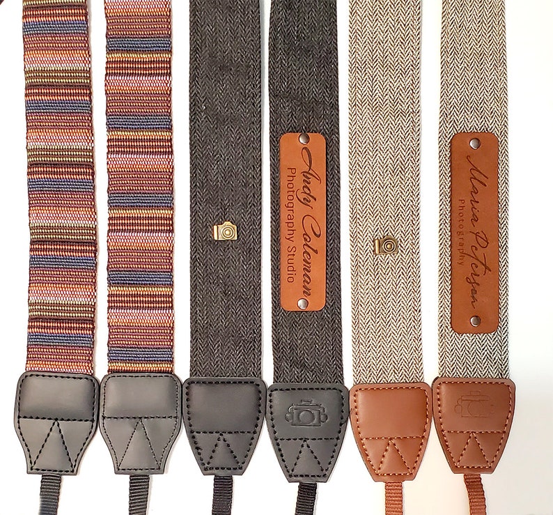 May include: Four camera straps with personalized leather name tags. The straps are made of different fabrics, including a striped fabric, a herringbone fabric, and a plain fabric. The name tags are brown leather with gold lettering.