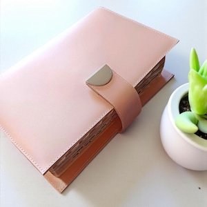 Op de afbeelding: Een lichtroze leren planner met een zilverkleurige sluiting. De planner is gesloten, vastgezet met een riem. De pagina's zijn bovenaan zichtbaar. Een kleine witte pot met een groene vetplant staat op de achtergrond.