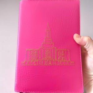 May include: A bright pink journal with a gold embroidered architectural design. The cover is a solid, vibrant pink with a textured appearance. A dark bookmark is visible. The journal is held by a hand.