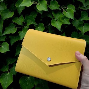 Op de afbeelding: Een mosterdgele clutch met een klepsluiting en een zilveren knoop. De clutch is gemaakt van een textuurrijk materiaal en wordt tegen een achtergrond van groene bladeren gehouden.