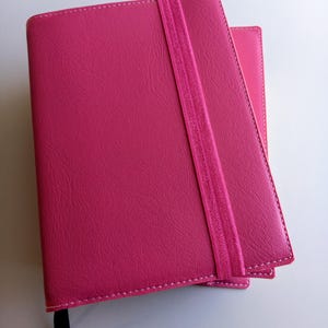 Op de afbeelding: Een roze lederen gebonden dagboek met een bijpassende elastische sluiting. Het dagboek heeft een gestikte rand en een zwart lint als bladwijzer.