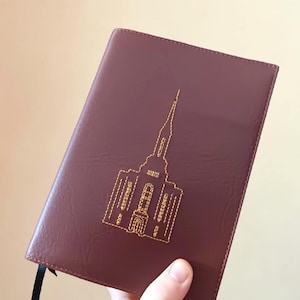 May include: A burgundy leather-bound journal with a gold embroidered church design. The journal has a black ribbon bookmark and is held at an angle. The cover is smooth and the stitching is detailed.