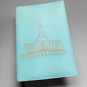 May include: A light blue journal with a gold embroidered building design. The cover is a solid light blue with a stitched border. A dark brown ribbon bookmark is visible. The journal is closed.