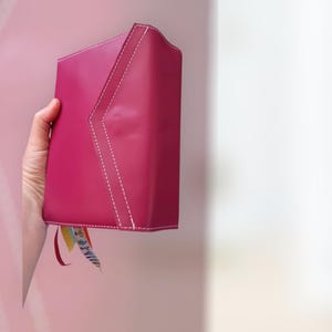 Könnte beinhalten: Ein magentafarbenes Lederjournal mit weißen Nähten und bunten Bändchen-Lesezeichen. Das Journal hat ein einzigartiges eckiges Design auf dem Einband. Das Journal wird in der Hand einer Person gehalten.