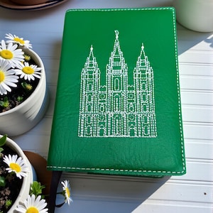 May include: Green journal with white stitching and a detailed white embroidered illustration of a building. The journal has a black ribbon bookmark. Daisies in a white pot are in the background.